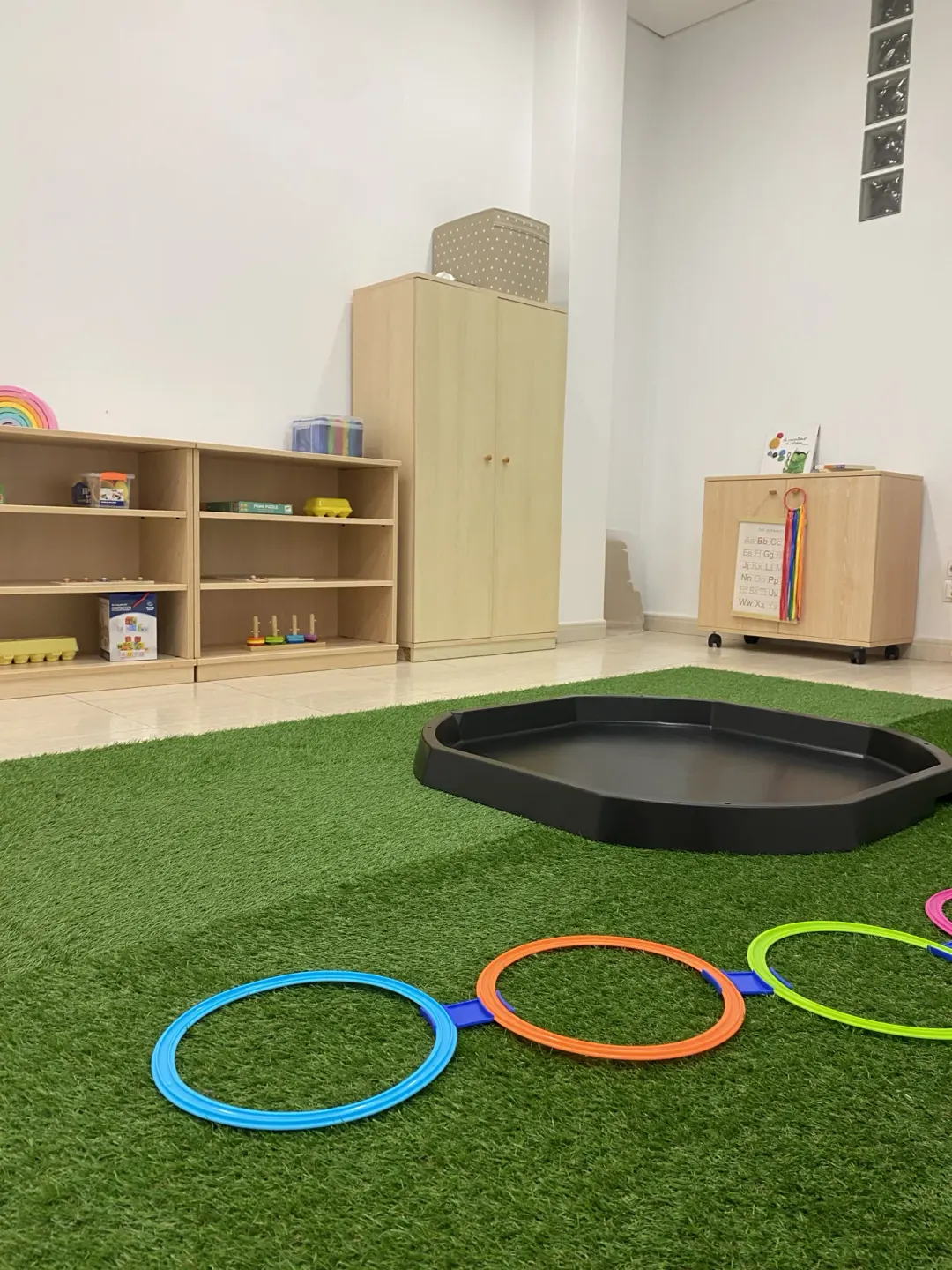 Sala de joc amb catifa verda, cèrcols de colors i prestatgeries amb materials.